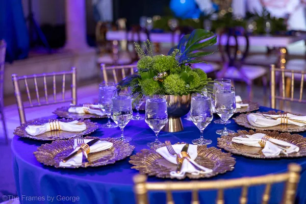 A formal event table setting with gold chiavari chairs, blue tablecloth, and floral centerpiece.