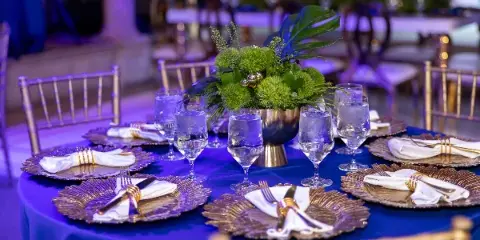 A formal event table setting with gold chiavari chairs, blue tablecloth, and floral centerpiece.