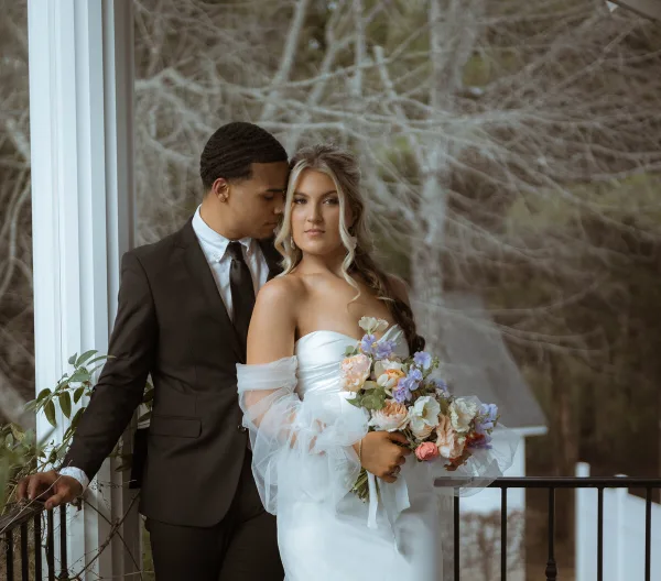A bride and groom posing together on a porch, with the bride holding a floral bouquet.