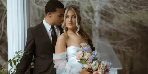 A bride and groom posing together on a porch, with the bride holding a floral bouquet.
