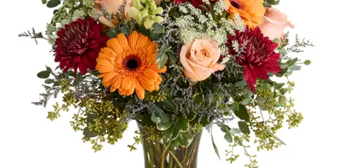 A floral arrangement featuring orange gerbera daisies, peach roses, and red dahlias in a clear glass vase.