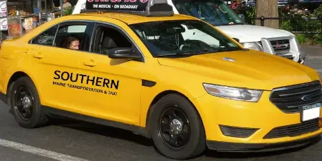 Yellow taxi sedan with 'Southern Maine Transportation & Taxi' branding driving on a city street