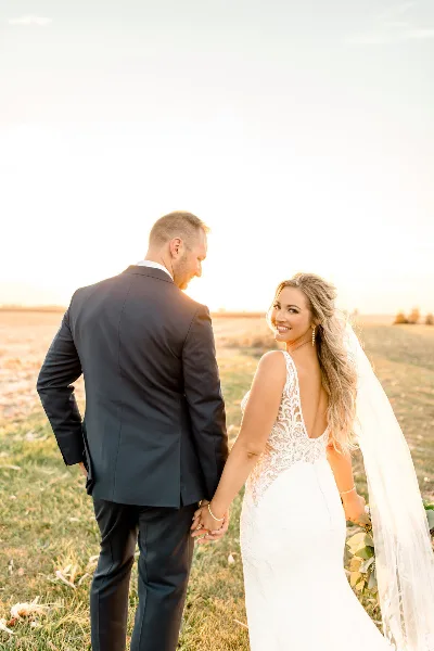 A bride and groom holding hands in a field at sunset, with the bride looking back at the camera.