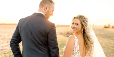 A bride and groom holding hands in a field at sunset, with the bride looking back at the camera.