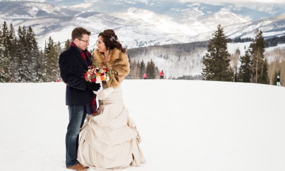 A bride and groom standing together in a snowy landscape with mountains in the background.