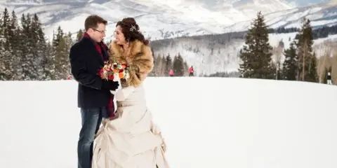 A bride and groom standing together in a snowy landscape with mountains in the background.
