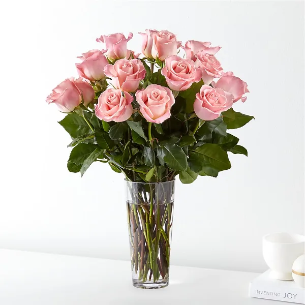 A bouquet of long-stemmed pink roses arranged in a clear glass vase on a white surface.