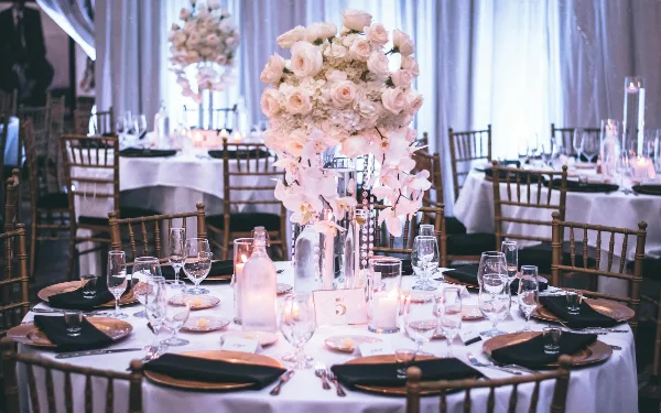 A wedding reception table set with white linens, gold chargers, black napkins, glassware, and a tall floral centerpiece, surrounded by wooden chairs.