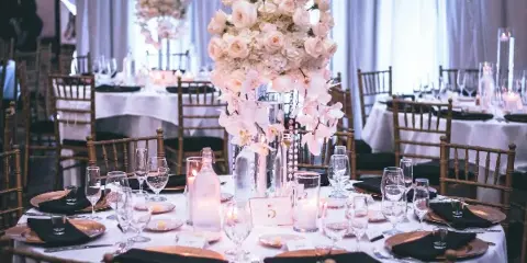 A wedding reception table set with white linens, gold chargers, black napkins, glassware, and a tall floral centerpiece, surrounded by wooden chairs.
