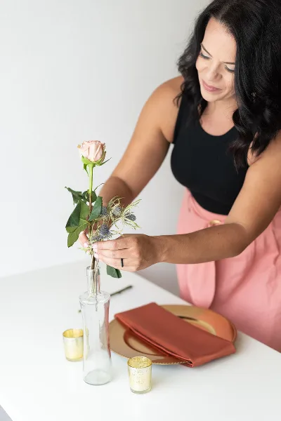 A woman arranging a pink rose and greenery into a glass vase on a table set with gold plates and candles.