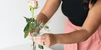A woman arranging a pink rose and greenery into a glass vase on a table set with gold plates and candles.