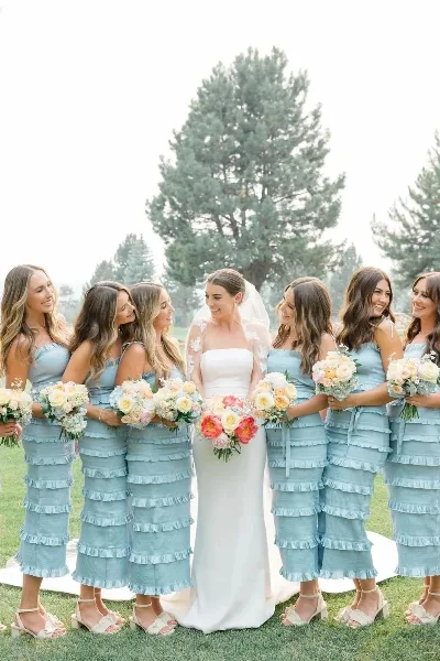 A bride in a white gown and veil stands with six bridesmaids in matching blue dresses, all holding bouquets of flowers outdoors on a lawn.