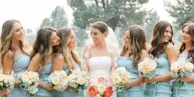 A bride in a white gown and veil stands with six bridesmaids in matching blue dresses, all holding bouquets of flowers outdoors on a lawn.