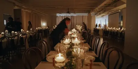 Interior view of a large event venue with long tables set with candles and floral centerpieces, with staff members arranging the space.