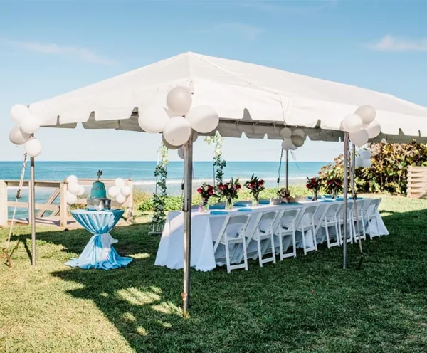 An outdoor event setup featuring a white tent, a long table with white linens and floral centerpieces, and white chairs arranged on a lawn near the ocean.