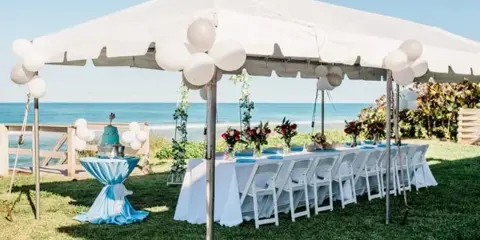 An outdoor event setup featuring a white tent, a long table with white linens and floral centerpieces, and white chairs arranged on a lawn near the ocean.