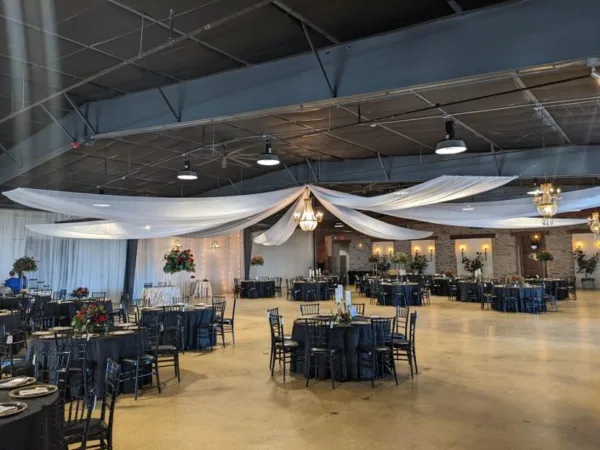 An indoor event venue featuring round tables, black chairs, and white fabric draping suspended from the ceiling.