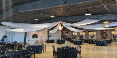An indoor event venue featuring round tables, black chairs, and white fabric draping suspended from the ceiling.