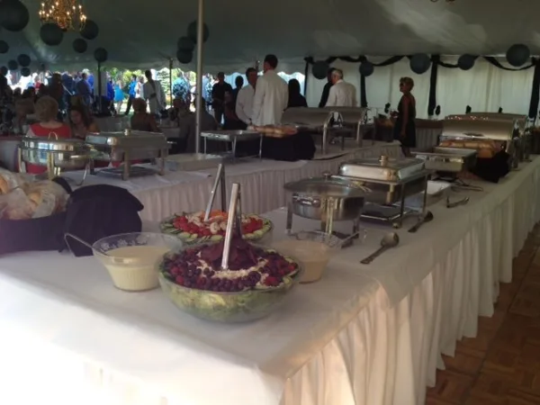 A long catering table set up under a tent with chafing dishes, food bowls, and serving utensils.