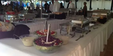 A long catering table set up under a tent with chafing dishes, food bowls, and serving utensils.