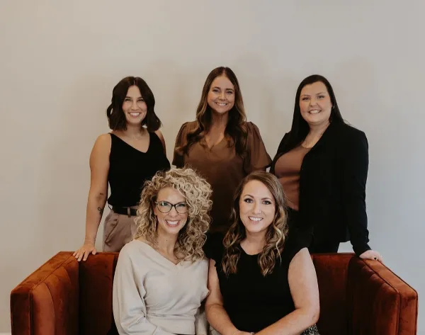 Group portrait of five smiling women, the team of White Dahlia Events, posing in front of a neutral background with a rust-colored velvet sofa.
