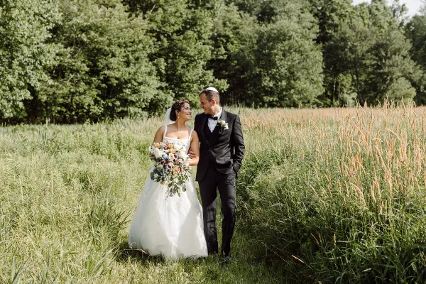 A bride and groom standing together in a grassy field, the bride holding a bouquet of flowers.