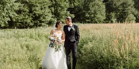 A bride and groom standing together in a grassy field, the bride holding a bouquet of flowers.