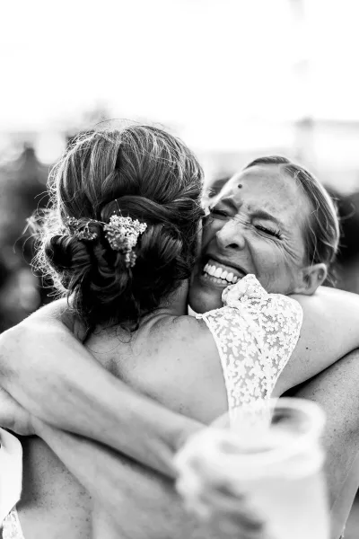 A black and white candid photograph of a bride in a lace dress laughing while hugging a guest.