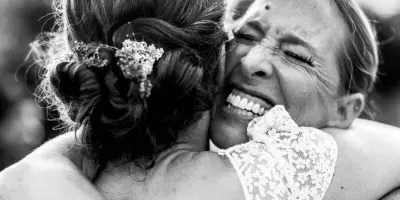 A black and white candid photograph of a bride in a lace dress laughing while hugging a guest.