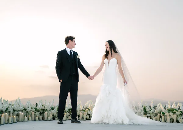 A bride and groom holding hands and smiling at each other in front of a floral and candle arrangement at sunset.
