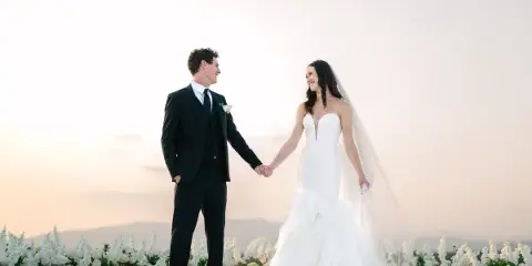 A bride and groom holding hands and smiling at each other in front of a floral and candle arrangement at sunset.