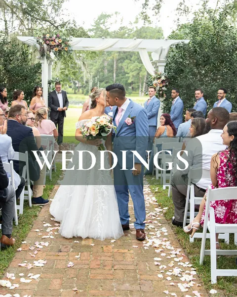 A bride and groom kissing under a white floral arch during an outdoor wedding ceremony with seated guests.