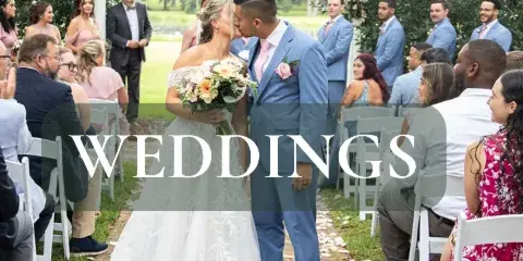A bride and groom kissing under a white floral arch during an outdoor wedding ceremony with seated guests.