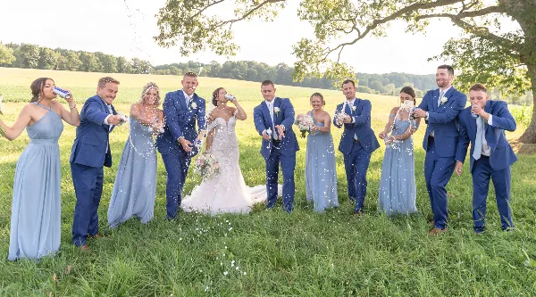 A wedding party, including the bride and groom, stands in a grassy field spraying champagne and celebrating.