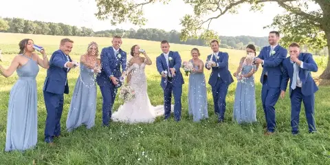 A wedding party, including the bride and groom, stands in a grassy field spraying champagne and celebrating.