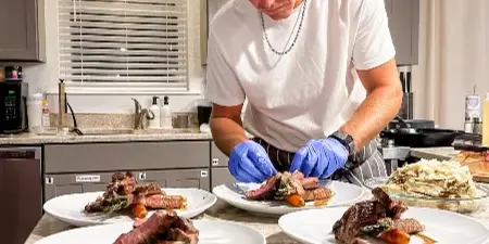 A chef wearing blue gloves plating a gourmet dish with sliced meat and vegetables on white plates in a kitchen.