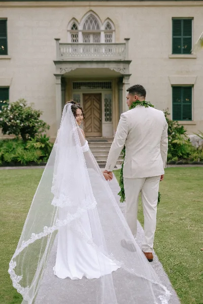 A bride and groom holding hands on a paved path in front of a large, elegant building with green shutters and manicured landscaping.