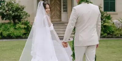 A bride and groom holding hands on a paved path in front of a large, elegant building with green shutters and manicured landscaping.