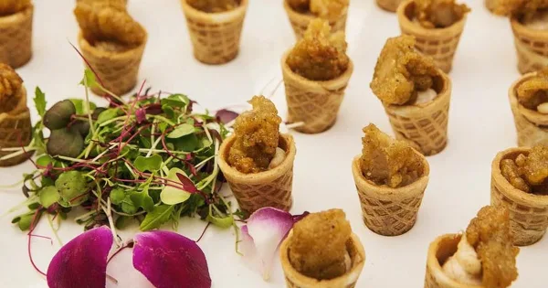 A close-up of a catering appetizer featuring waffle cone bites filled with savory ingredients, garnished with fresh microgreens and purple flower petals on a white surface.