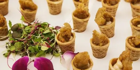 A close-up of a catering appetizer featuring waffle cone bites filled with savory ingredients, garnished with fresh microgreens and purple flower petals on a white surface.