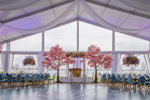 Interior view of a white wedding pavilion with a dance floor, blue chairs, and floral arrangements, overlooking a body of water.