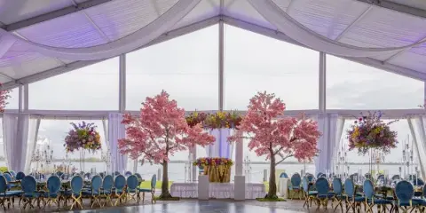 Interior view of a white wedding pavilion with a dance floor, blue chairs, and floral arrangements, overlooking a body of water.