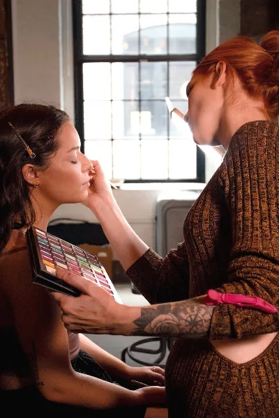 A makeup artist applying makeup to a client's face while holding a large eyeshadow palette.
