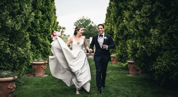A bride and groom walking hand-in-hand through a garden path lined with tall green hedges.