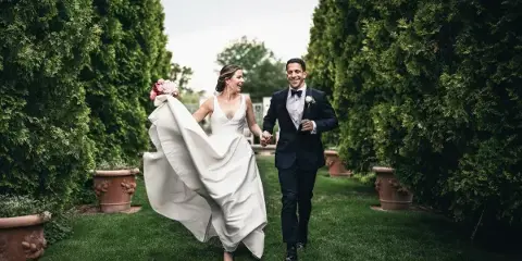 A bride and groom walking hand-in-hand through a garden path lined with tall green hedges.