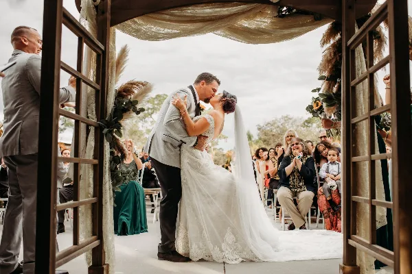 A bride and groom share a kiss under a wooden archway decorated with pampas grass and greenery during an outdoor wedding ceremony.