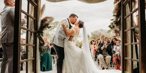 A bride and groom share a kiss under a wooden archway decorated with pampas grass and greenery during an outdoor wedding ceremony.