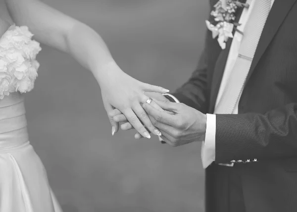 A black and white close-up of a bride and groom holding hands, with the groom placing a wedding ring on the bride's finger.