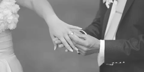 A black and white close-up of a bride and groom holding hands, with the groom placing a wedding ring on the bride's finger.