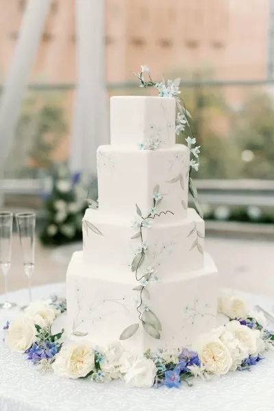 A four-tier white wedding cake decorated with hand-painted floral vines and fresh flowers, displayed on a table with champagne flutes.